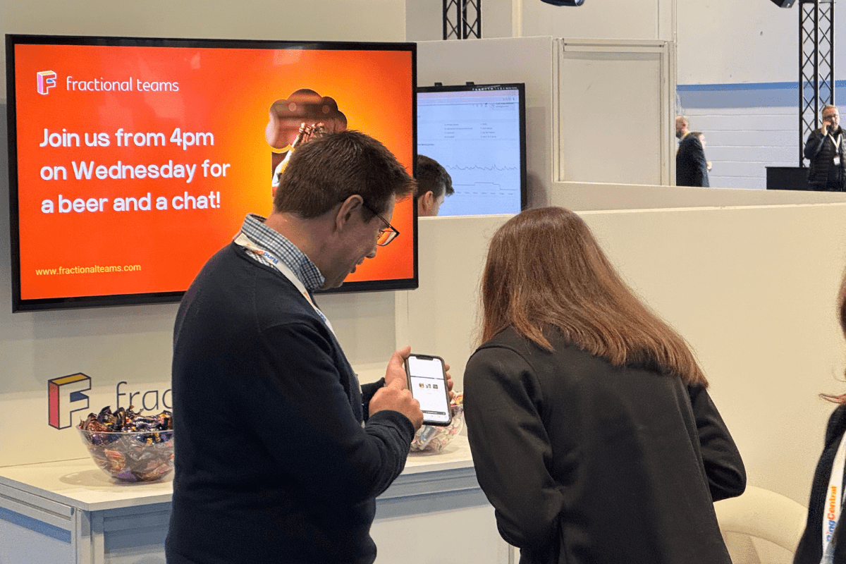 Two people stand at a booth in a convention hall. A man shows something on his phone to a woman. Behind them, a screen displays an invitation for a Wednesday event. Bowls of candy are visible on the counter.