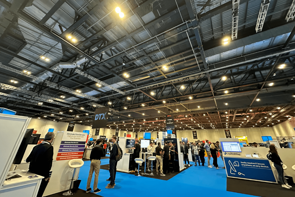 In the large, brightly lit exhibition hall, people gather around booths showcasing digital technology solutions. Attendees engage with exhibitors. Company signs and screens displaying information create a buzz of excitement and innovation.